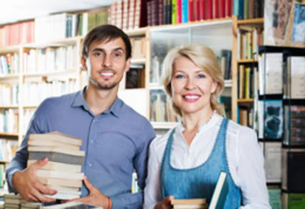 Зрелая женщина и молодой мужчина стоят в библиотеке с книгами в руках